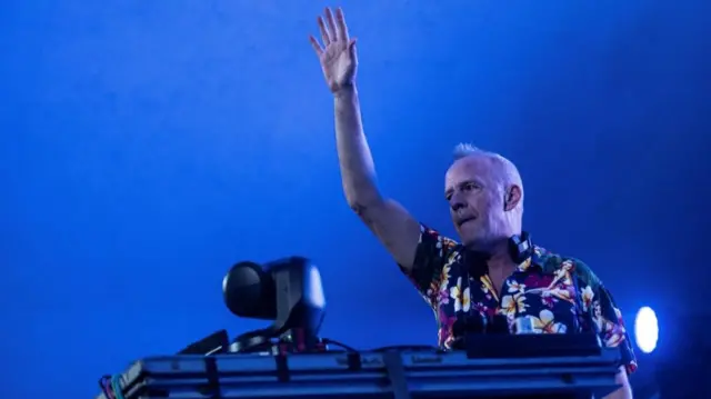 Fatboy Slim stands behind DJ decks in front of a blue background