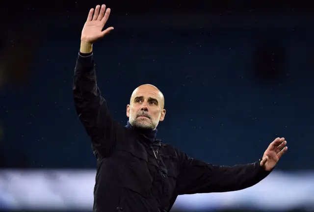 Pep Guardiola, a bald man with a salt and pepper goatee beard, wears a black jacket. He is is waving towards fans in the distance, in front of the camera, in a photograph taken at night and illuminated by stadium floodlights