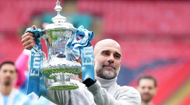 Pep Guardiola, a bald man with a salt-and-pepper goatee beard, smiles in a stadium while holding the silver FA Cup trophy adorned in sky blue and white ribbons. Man City players are in the background, out of focus