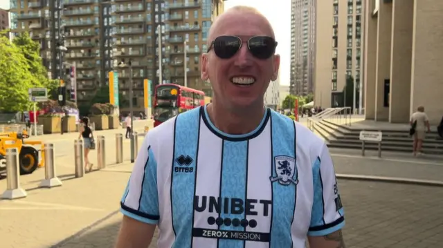 Boro fan Mark on Wembley Way in London. He is wearing a Middlesbrough shirt with white and pale blue stripes with the club crest n black and white and the word Unibet in black. He is wearing sunglasses and has a big smile on his face. In the background is a yellow digger and rows of flats are behind him
