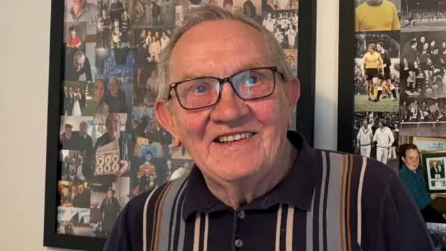 A man in his 80s with short grey hair smiles for the camera. Behind him is a framed compilation of photographs from his football-playing days.