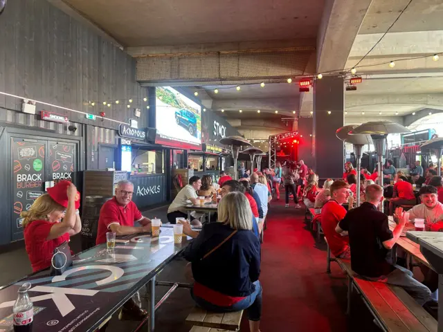 A wide view of the Stack in Middlesbrough with rows of tables and Middlesbrough fans sitting at them, many of them wearing red and white Middlesbrough shirts. There is a big television screen on the wall on the left hand side