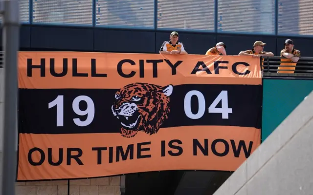 Four Hull fans standing next to a wall with a huge Hull flag draped over the side. it has the words Hull City AFC our time is now and 1904 in the middle surrounding a tiger's head