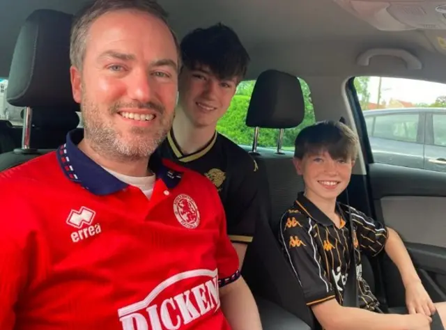 A man wearing a red Middlesbrough top is in a car with his two young sons both wearing Hull City shirts.