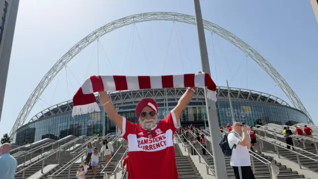A Boro fan in front of Wembley stadium. The white arch and the stadium are behind him and he is standing in front of the steps. He is holding a red and white Middlesbrough scarf above his head. He is wearing a red and white Middlesbrough shirt with Ramsdens Currency written in red on a white band across the middle of the shirt. He has a red and white turban on and is wearing sunglasses and has a white beard