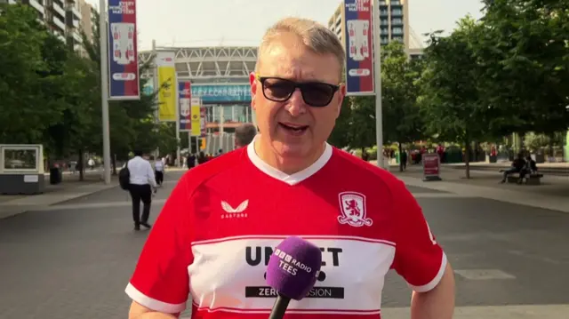 A Middlesbrough fan stands on Wembley Way. He has blond hair and sunglasses. He is wearing a red and white Middlesbrough shirt and is holding a purple microphone with BBC Tees on it. The stadium is behind him in the distance