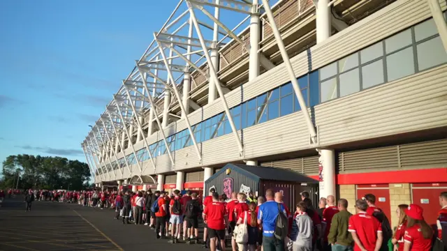 A queue of Middlesbrough fans outside the Riverside Starium in Middlesbrough. Many of them are wearing red and white Middlesbrough shirts and are standing in front of the ground which rises above them and is a white building with white supports coming out of the side