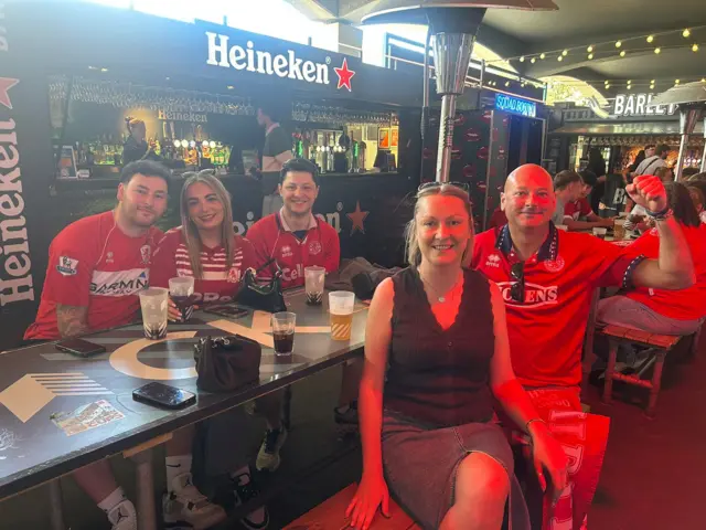 Middlesbrough fans sitting around a table. A woman sits between two men all wearing Middlesbrough shirts at the back and at the front are a man and a woman - the man is also wearing a Boro shirt and the woman is wearing a black sleeveless top and a denim skirt. Behind them is a bar