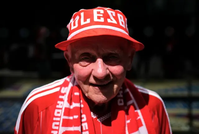 An older male Boro fan wearing a red and white bucket hat with Middlesbrough written around it and wearing a red and white Boro hat and and a red and white Boro scarf