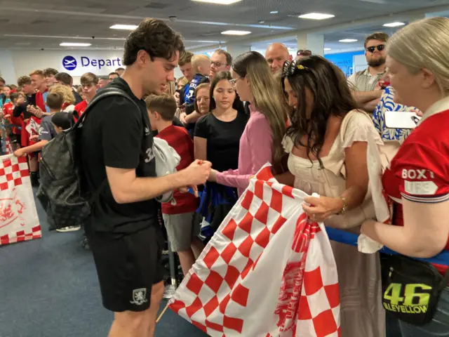 Crowds of three deep stand behind a barrier in the airport to greet Middlesbrough players. One woman wearing a white dress is getting her red and white flag signed by player Hayden Hackney, who is wearing a black shirt and shorts. There is a sign on the wall behind them that reads: "Departures."