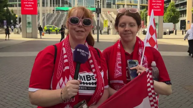 Two women wearing red and white Middlesbrough shirts. The lady on the left has sunglasses on and has long red hair. She is wearing a Middlesbrough scarf and is holding a purple microphone with BBC Tees on it. The lady on the right is wearing sunglasses on her head and has a scarf around her neck and is holding a Middlesbrough flag with red and white squares over her shoulder and has another flag on her other hand