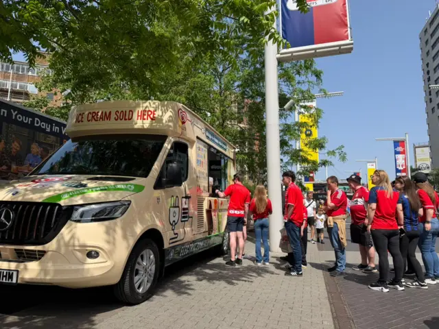 A queue of Boro fans in red and white shirts queuing for ice creams. The van is a cream colour and has ice cream sold here in red letters on the front of the van