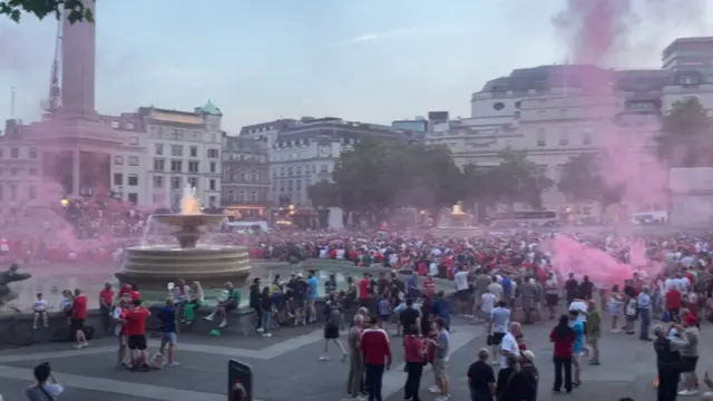 A wide shot of Trafalgar Square with one of the fountains and the base of Nelson's column on the left hand side of the photo and white coloured buildings at the back of the image surrounding the square. There aer hundreds of people, many of them wearing red Middlesbrough shirts. There is red smoke from flares drifting across the square