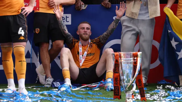 Oli McBurnie celebrates with his hands raised while sat on the floor behind the play-off final trophy