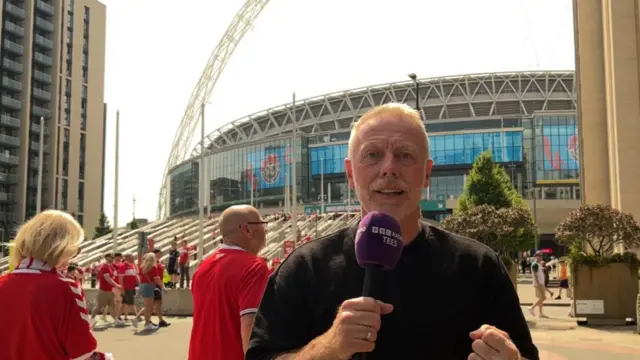 Craig Hignett is wearing a black t-shirt and is holding a purple microphone with the BBC Tees logo on it in white. His first on his other hand is clenched. He is standing in front of Wembley Stadium with the part of the arch