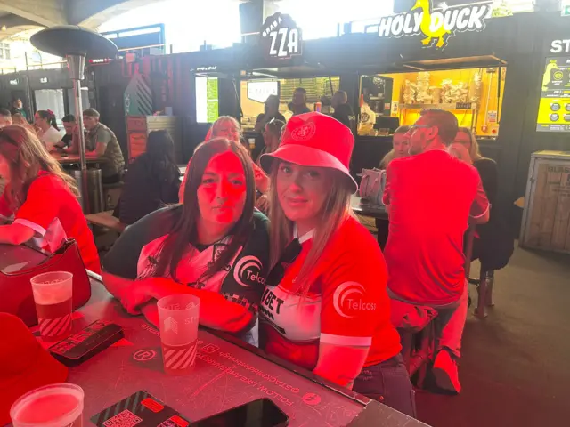 Two female Middlesbrough fans sitting at a bar with pints of beer in front of them. The fan on the left has long brown hair and is wearing a green and white Middlesbrough top. The fan on the right is wearing a red Middlesbrough bucket hat and a red and white Middlesbrough top