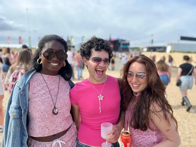 Kameel, Emmanuel and Esther posing to the camera in the sun. Kameel is smiling an wearing earrings. She has a denim jacket over a pink top and a diamond shaped necklace. Emmanuel is wearing a pink top and wearing a star necklace. Esther is wearing a pink patterned vest. They are all wearing sunglasses. Festival-goers can be seen in the background.
