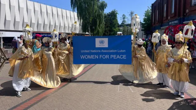 Six women dressed in gold and white costumes hold lanterns and a sign that says: United Nations Association Luton - Women for Peace