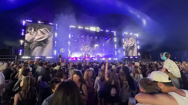 Crowd of people in festival tent in front of stage and big festival screens. The stage lights are purple.
