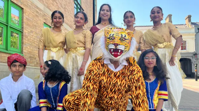 Four girls stand at the back with gold costumes, flanking Blenda Shibu, who is wearing a red and gold sari and gold choker. There is a buy wearing a red turban and white shirt kneeling at the front, with two girls in bright blue costumes. They are kneeling next to a person dressed in a tiger costume.