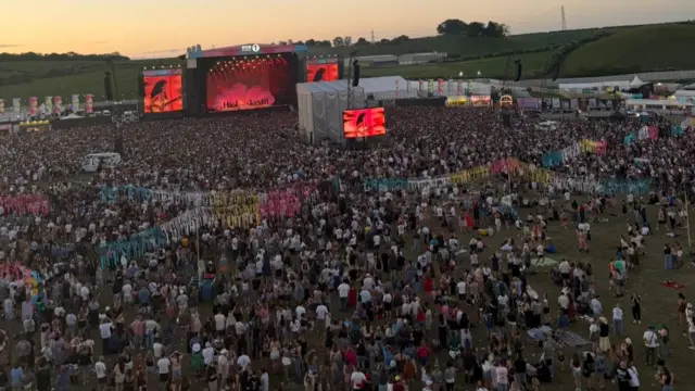 Overhead shot of festival with the sun going down. Lots of people facing main stage.