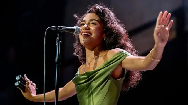 Olivia Dean wears a green satin off-the-shoulder dress. She has brown curly hair which is cascading down her back. She has her arm raised out on either side of her.