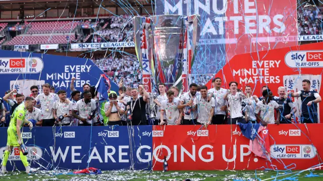 Bolton players celebrate with the trophy, confetti in front of a 'We are going up' sign