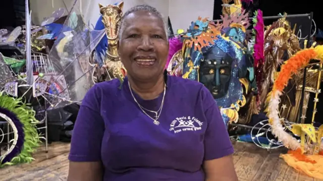 Margaret Matthew is wearing a putple T-shirt with a ST Kitts, Nevis & Friends logo. She is sitting in a room full of carnival costumes.