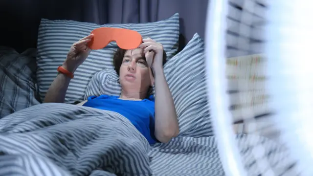 Woman is using eye mask in bed while electric fan is blowing in the foreground