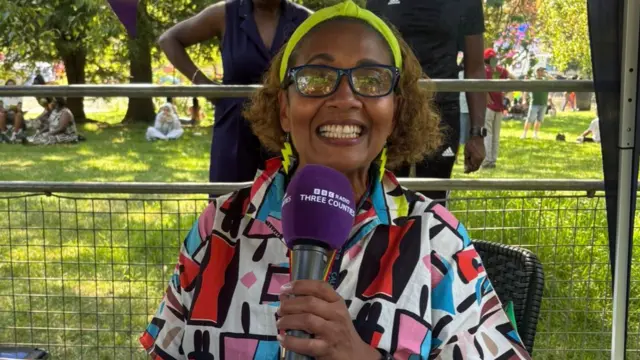 Babs Michel is grinning at the camera and holding a purple microphone