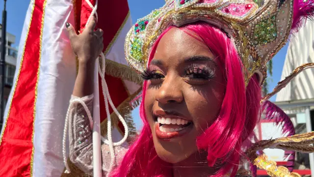 Amber Amber Ogunsanya-William is dressed in a pink wig and is looking directly at the camera. She is smiling and is wearing an elaborate headpiece and is holding a flag.
