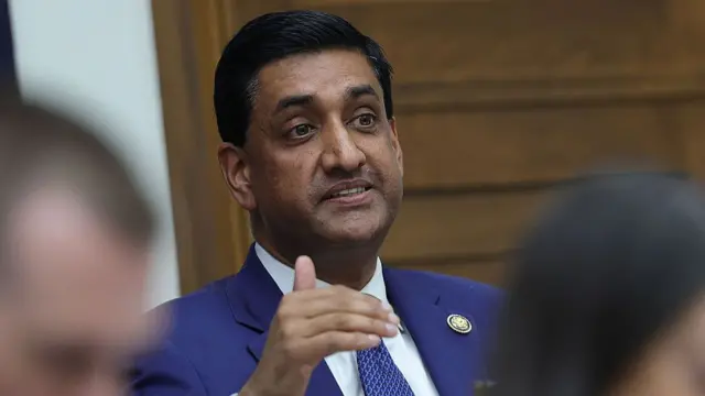 Democratic Congressman Ro Khanna in a dark blue suit, white shirt and patterned blue tie speaking. There's a wooden door behind him to his left