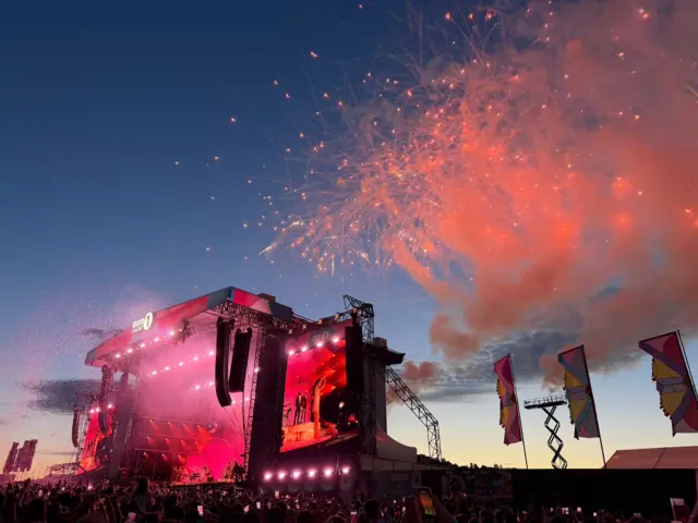 Fireworks explode to the right of the stage with a big screen next to the stage showing Olivia Dean waving to the crowd. Everything is bathed in pink light
