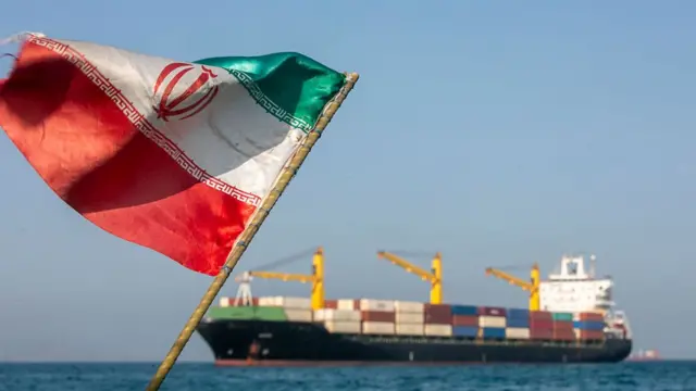 A commercial ship carrying containers across the Strait of Hormuz sailing on water, as an Iranian flag waves in the foreground