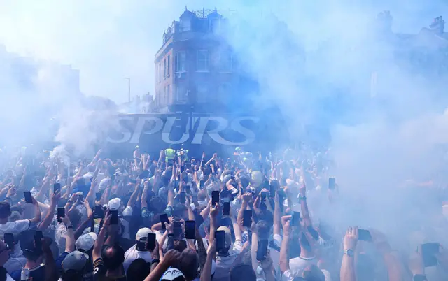 Tottenham supporters hold up their phones and there is smoke as the Tottenham bus is in the background
