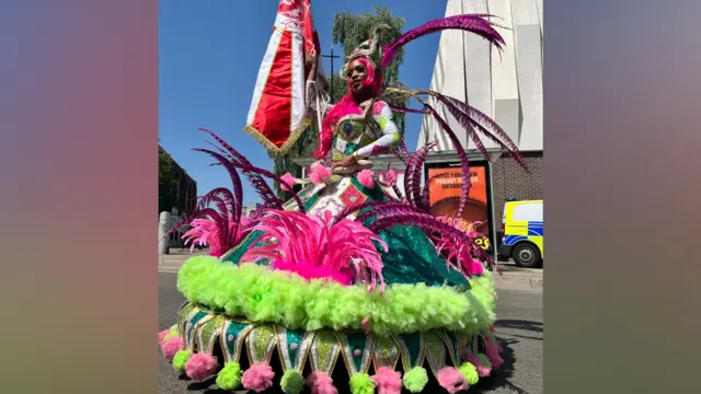A woman in an extravagant pink, green and white outfit with a wide hooped skirt, and feathers coming off it, holds a red and white flag.