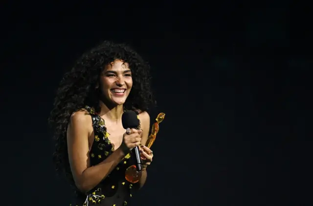 Olivia Dean wearing a black dress with yellow flowers and dots on it. She is holding a microphone and her artist of the year award form the Brit Awards