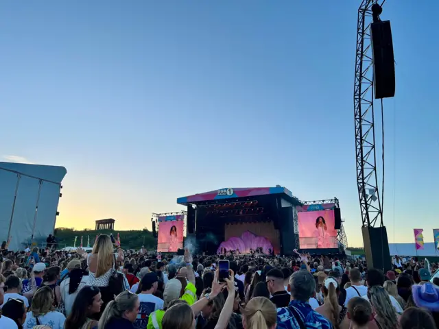 Crowd of people looking at festival stage with large festival screens. Olivia Dean in a pink dress is on the screens.The sky is turning darker but still blue. It is turning to dusk.
