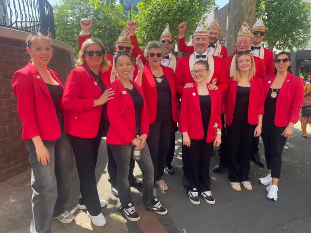 A group of men, women and girls, dressed in red jackets and black tops and trousers, with some wearer patterned hats, smile at the camera. Some of the men are punching the air.