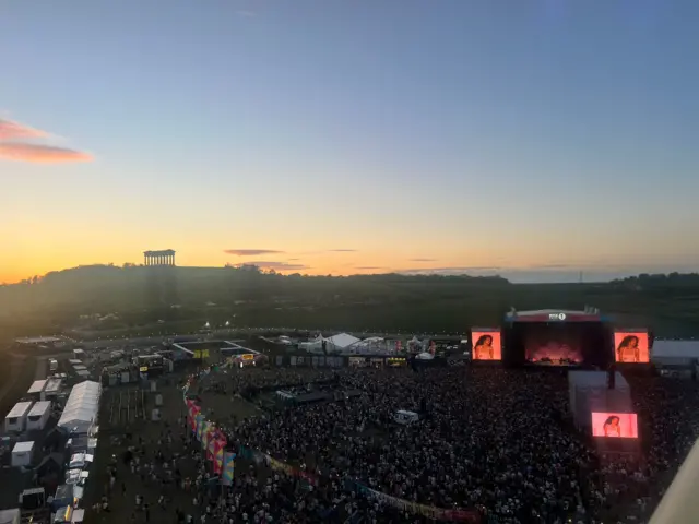 Overhead shot of festival with the sun going down. Lots of people facing main stage.