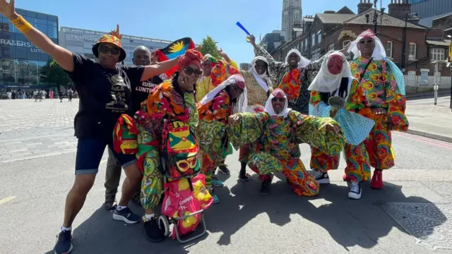A group of people pose for the camera with their arms out wearing brightly-patterned tops and trousers and white headscarves.