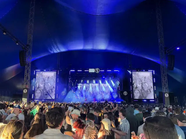 Large crowd of people in a large festival tent with a stage and big festival screens in the front. The inside of the tent is dark.