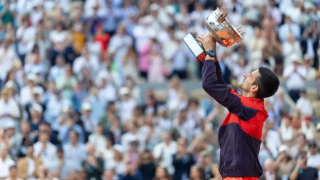 Novak Djokovic, wearing an orange and dark blue tracksuit top, stands side on and raises the silver trophy in two hands above his head with the crowd blurred in the background