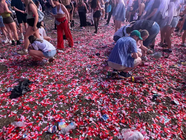 Lots of red and pink confetti on the floor with people in festival walking and crouching among it.