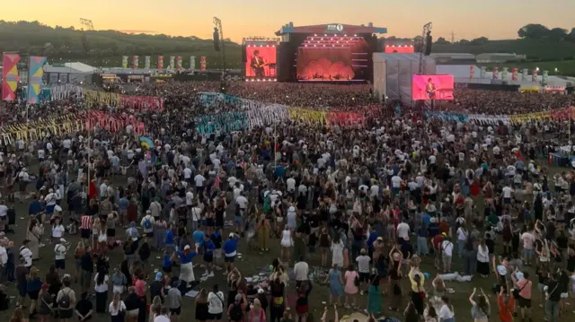 Overhead shot of festival crowd watching Olivia Dean as the sun goes down. Thousands of thousands of people are in the shot. Pink light comes from the stage.