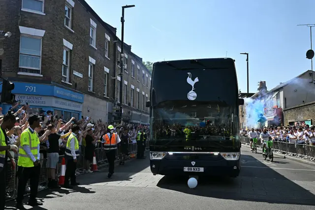 A bus with a Tottenham logo on the window driving down a road with supporters lining the streets