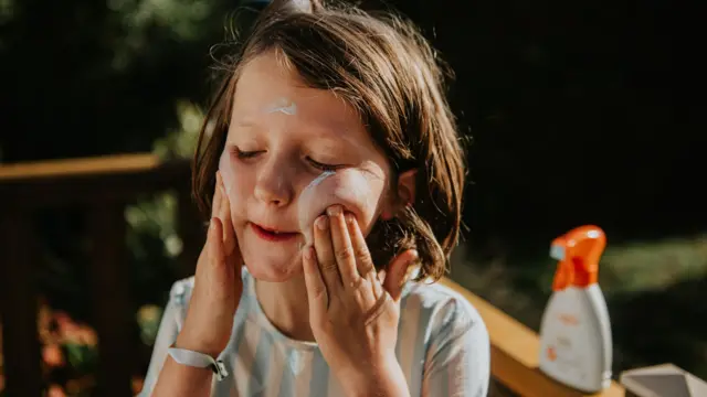 A child is applying sunscreen on her cheeks while standing outside in a backyard