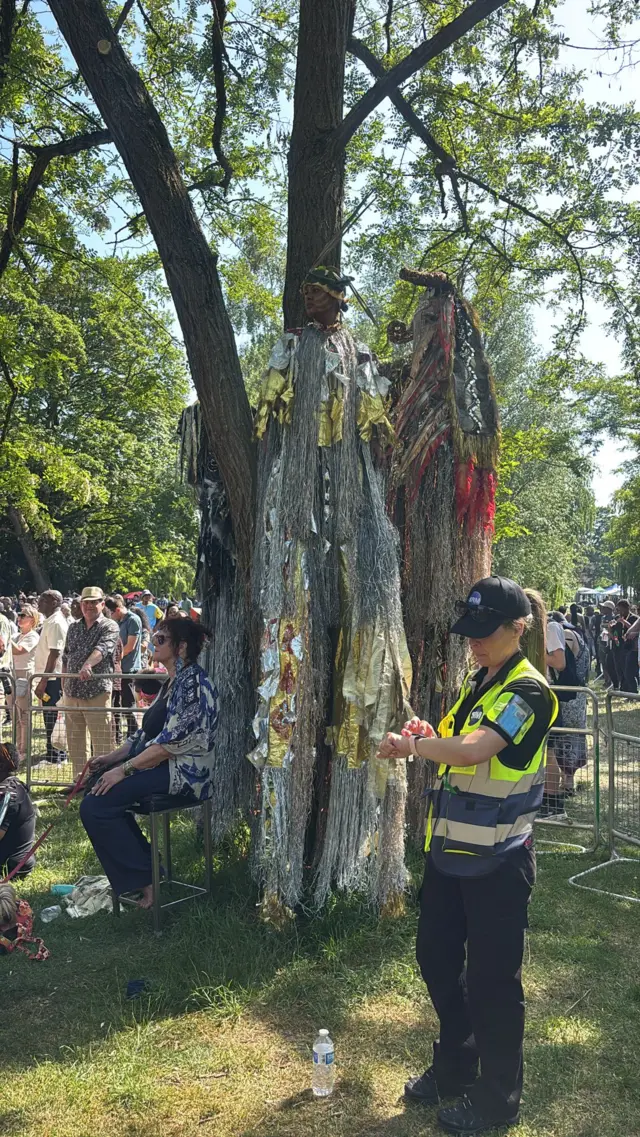 People standing on stilts in costumes with strings of material sheltering under a tree in a park