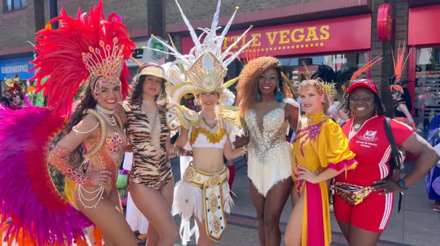 A group of six women wear elaborate and brightly-coloured costumes and pose in front of a shop.
