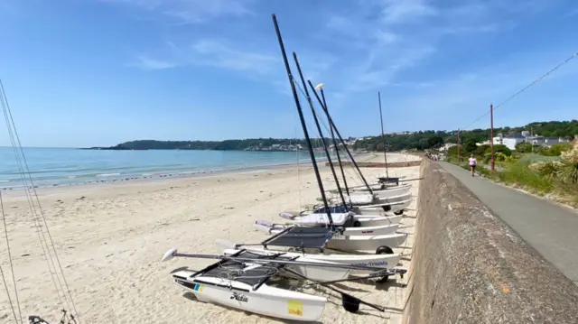 A row of small sailing boats sits on a sandy beach beside a coastal path, with calm blue sea, a distant headland and scattered houses under a bright, lightly clouded sky.